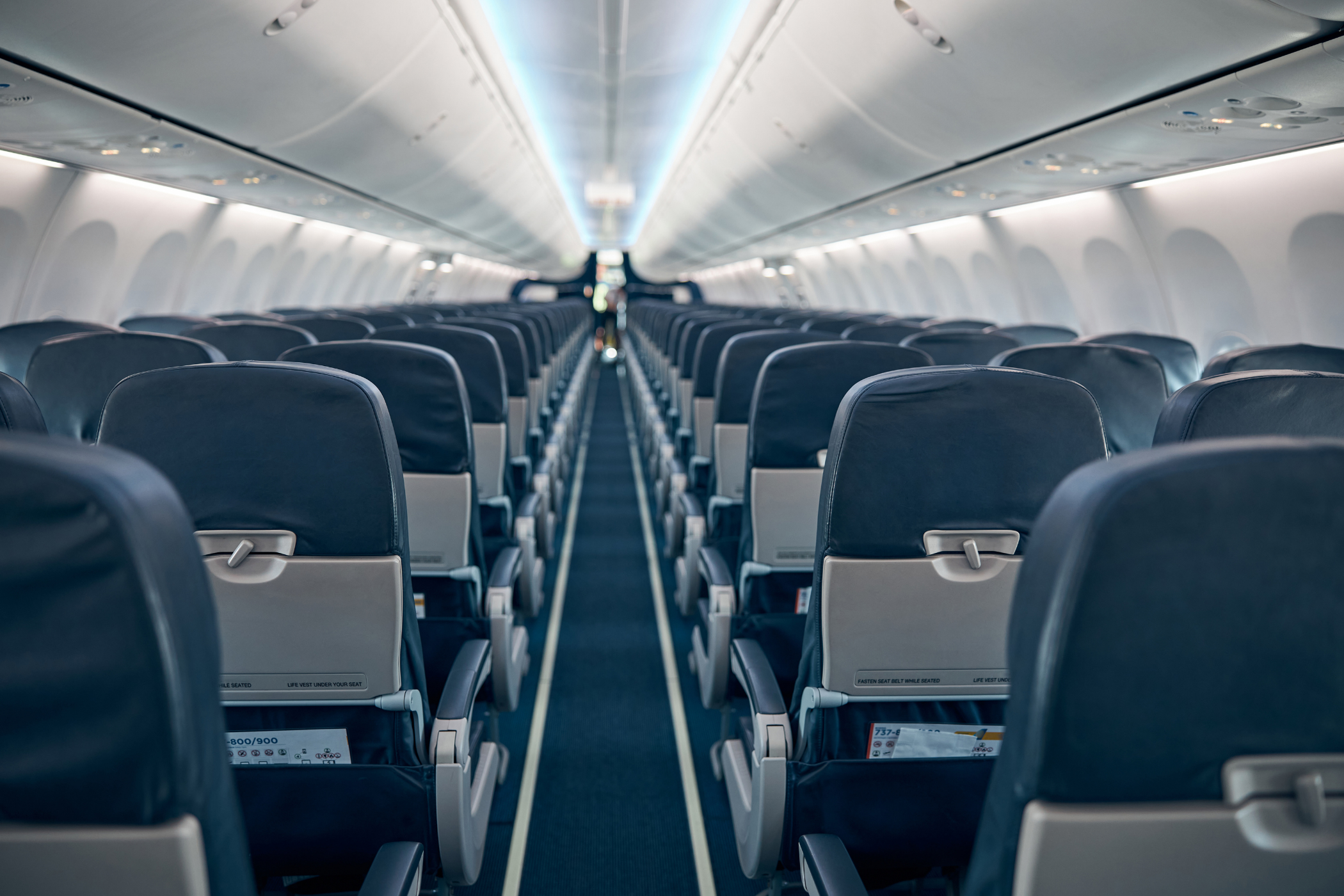 Interior view of an empty airplane cabin with rows of black seats and an aisle running down the middle.