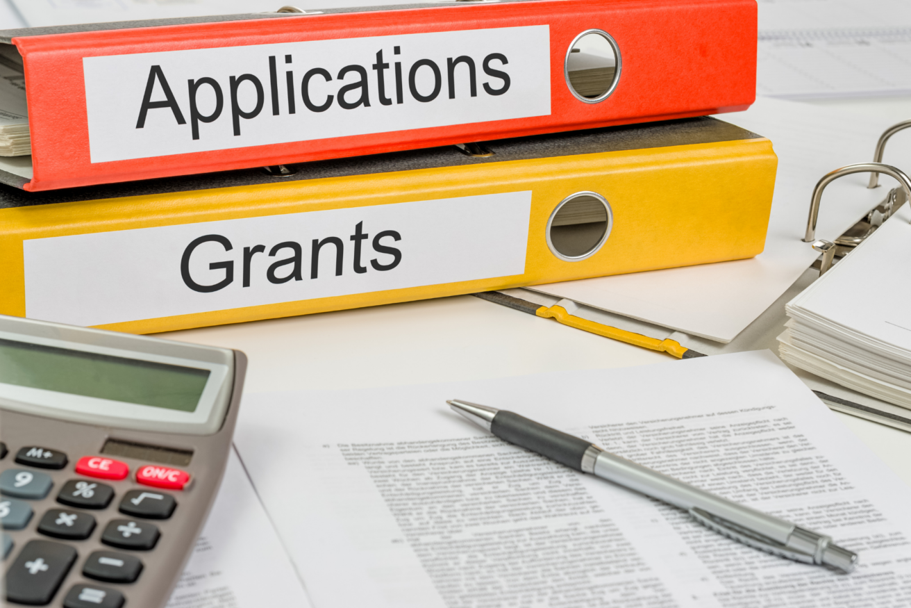 Two labeled ring binders, one red for "Applications" and one yellow for "Grants," on a desk with documents, a pen, and a calculator.