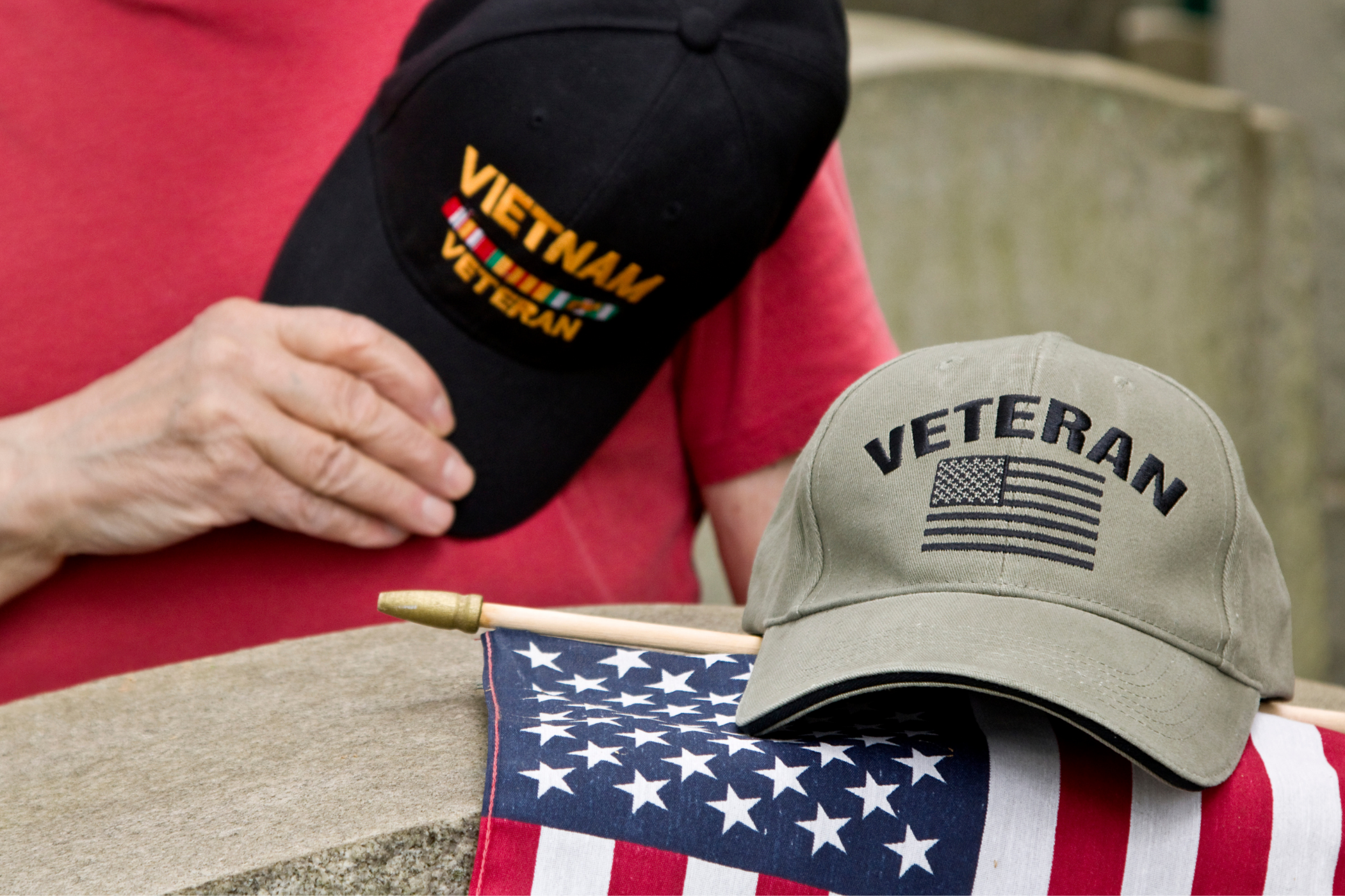 Person in red shirt holding a black "Vietnam Veteran" cap, with a grey "Veteran" cap and an American flag placed on a grave marker.