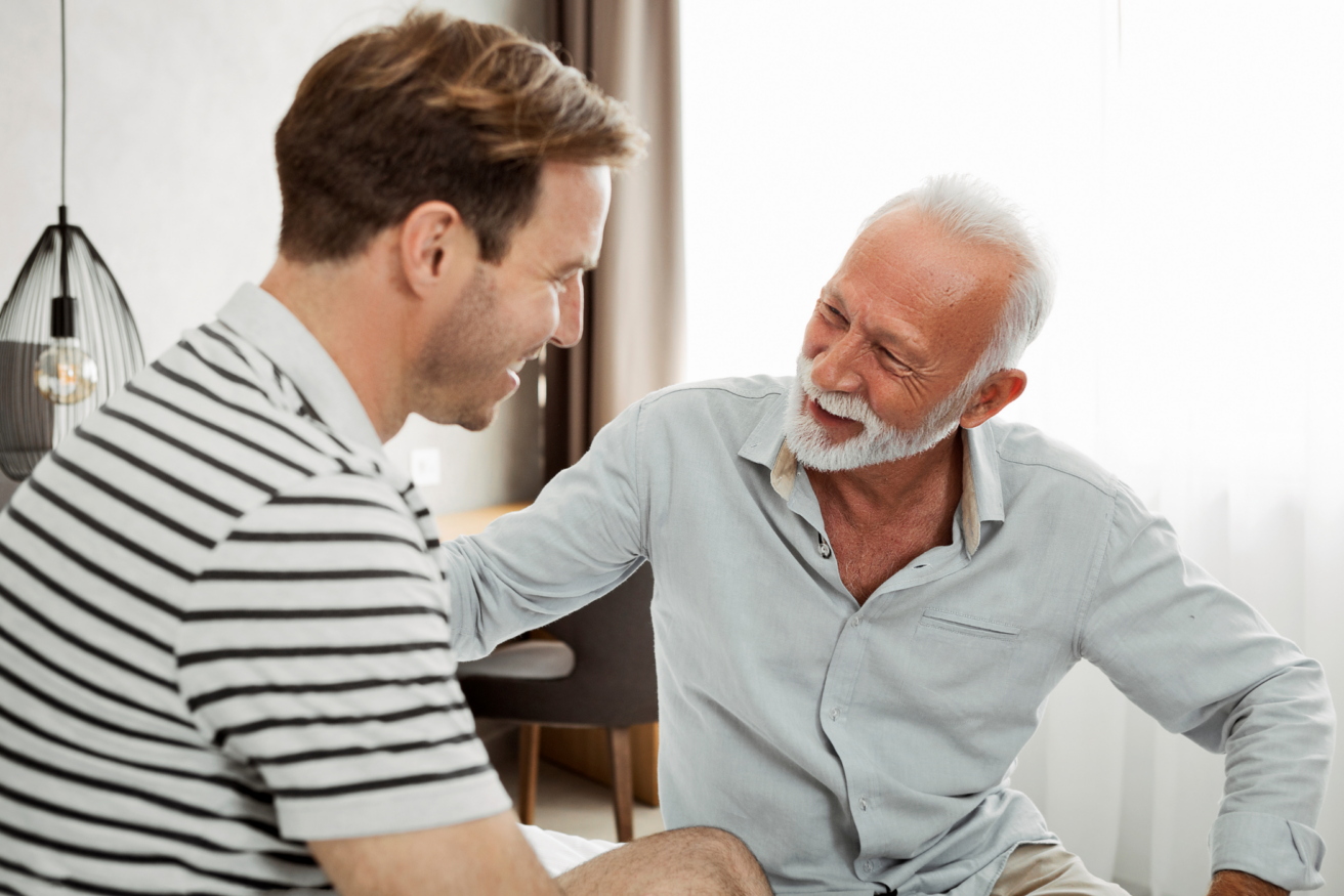 Two men, one older with white hair and a beard, the other younger with brown hair, are seated and smiling at each other in a brightly lit room.