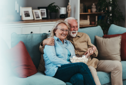 Elderly couple sitting on a couch, smiling while sharing a bowl of popcorn in a cozy living room setting with shelves and framed pictures in the background.