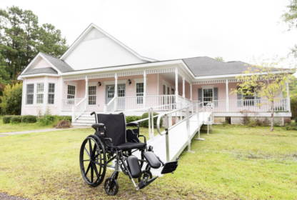 A wheelchair rests on the lawn facing a house with a wheelchair-accessible ramp leading up to the front porch.