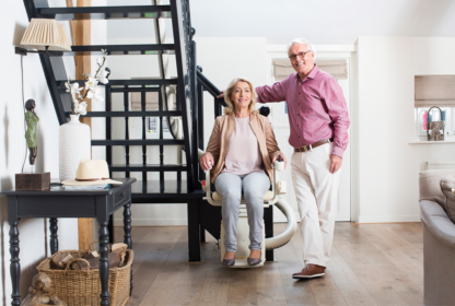 An elderly woman sits on a stairlift at the bottom of a staircase while an elderly man stands beside her in a bright, modern living room.