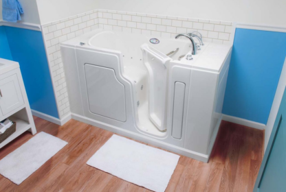 A white walk-in bathtub with a door and built-in seat in a bathroom with blue and white walls and wooden flooring. Two white bath mats are placed in front of the tub.