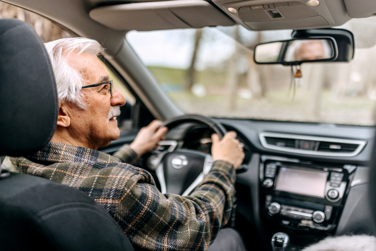 Older adult man with gray hair and glasses sits in the driver's seat of a car, hands on the steering wheel, looking forward through the windshield.