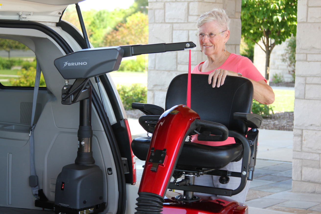 A woman wearing a pink shirt uses a mechanical lift to load a red mobility scooter into the back of a vehicle.