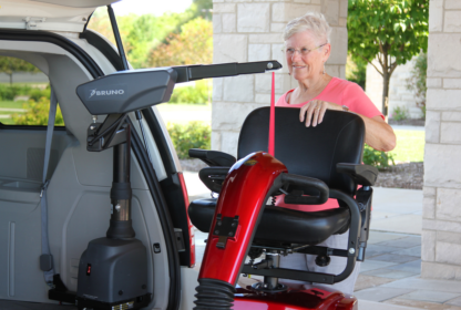 A woman wearing a pink shirt uses a mechanical lift to load a red mobility scooter into the back of a vehicle.