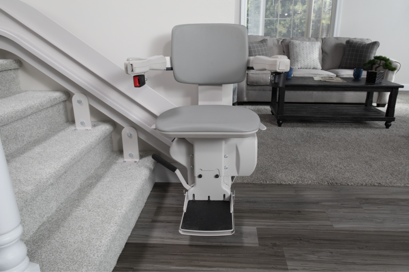 A stair lift chair installed on the side of a staircase leading to a carpeted living room with a gray sofa and a coffee table.