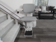 A stair lift chair installed on the side of a staircase leading to a carpeted living room with a gray sofa and a coffee table.