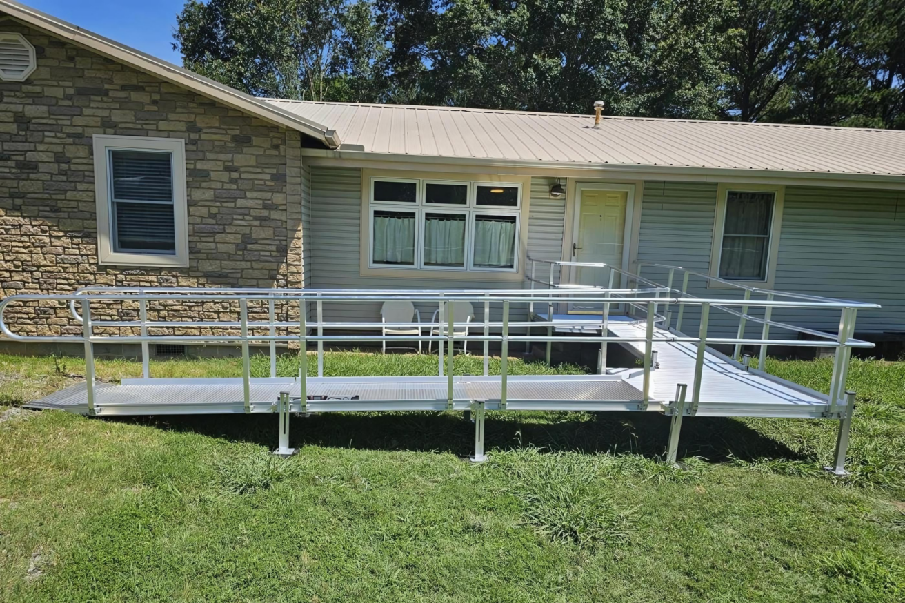 A metal wheelchair ramp installed outside a single-story house with a stone and siding exterior, leading to a door at the back.