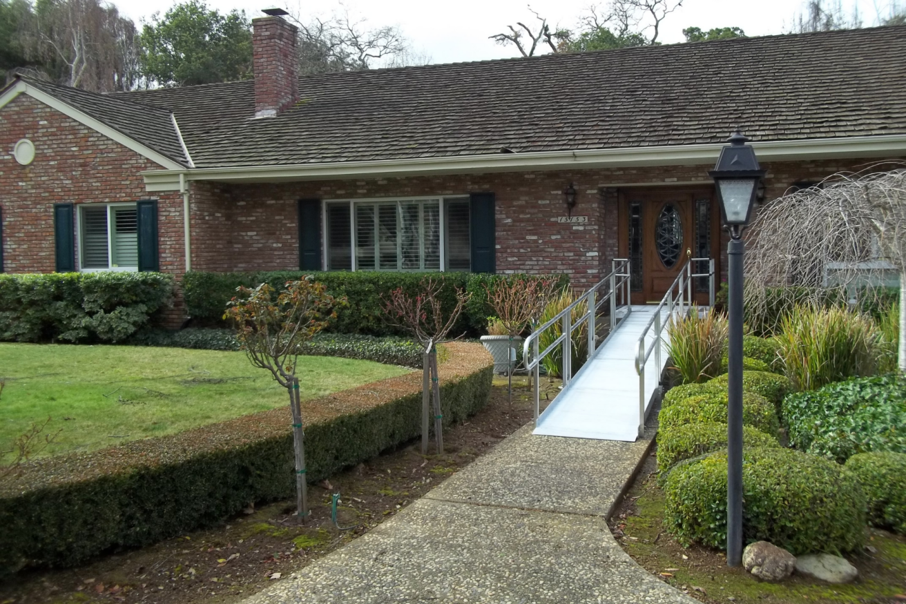 A one-story brick house with a shingled roof has a ramp leading to the front door. The yard includes trimmed bushes and a lamp post along a walkway.