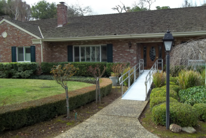 A one-story brick house with a shingled roof has a ramp leading to the front door. The yard includes trimmed bushes and a lamp post along a walkway.