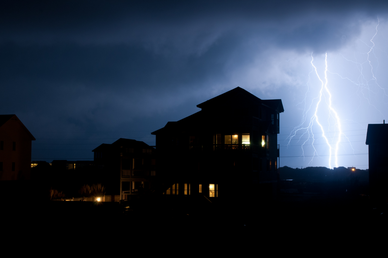 Silhouette of houses against a dark sky illuminated by a striking bolt of lightning.