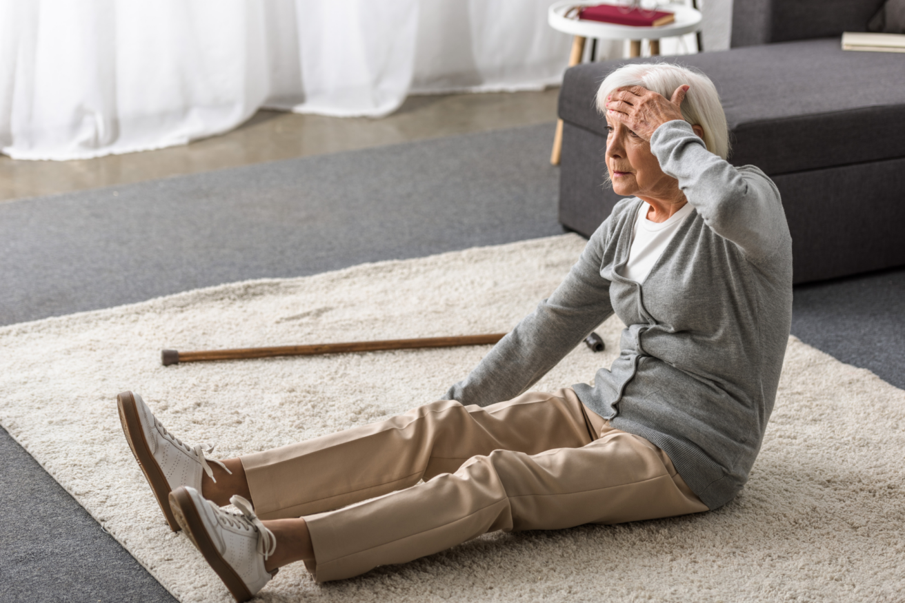 An older woman sitting on a rug looks distressed, holding her head with one hand. A wooden cane is lying on the carpet next to her.