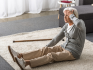 An older woman sitting on a rug looks distressed, holding her head with one hand. A wooden cane is lying on the carpet next to her.