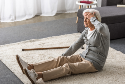 An older woman sitting on a rug looks distressed, holding her head with one hand. A wooden cane is lying on the carpet next to her.