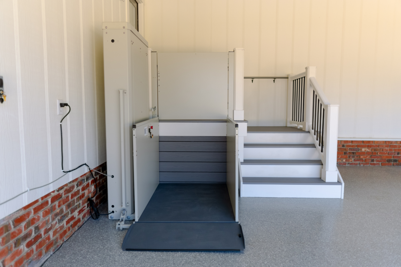 A residential home elevator lift positioned next to a small set of stairs. The lift has safety railings and is fully lowered, ready for use. The surrounding area has white walls and a brick accent.