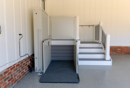 A residential home elevator lift positioned next to a small set of stairs. The lift has safety railings and is fully lowered, ready for use. The surrounding area has white walls and a brick accent.