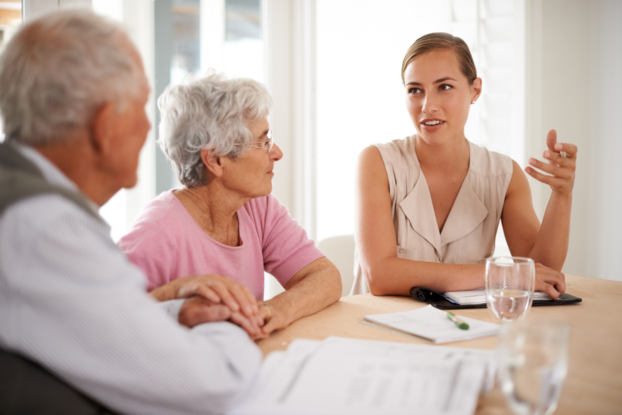 A young woman is talking to an elderly couple at a table with documents, a notepad, and a glass of water.