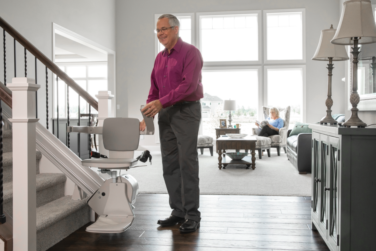 A man in a purple shirt stands by a stairlift at the bottom of a staircase. A person is seated on a couch in the background. The room is bright and well-lit with large windows.