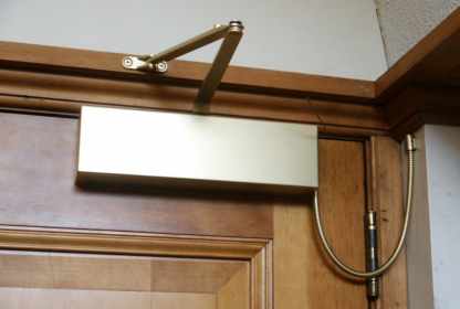 A brass door closer mounted at the top of a wooden door, equipped with an arm and chain mechanism.