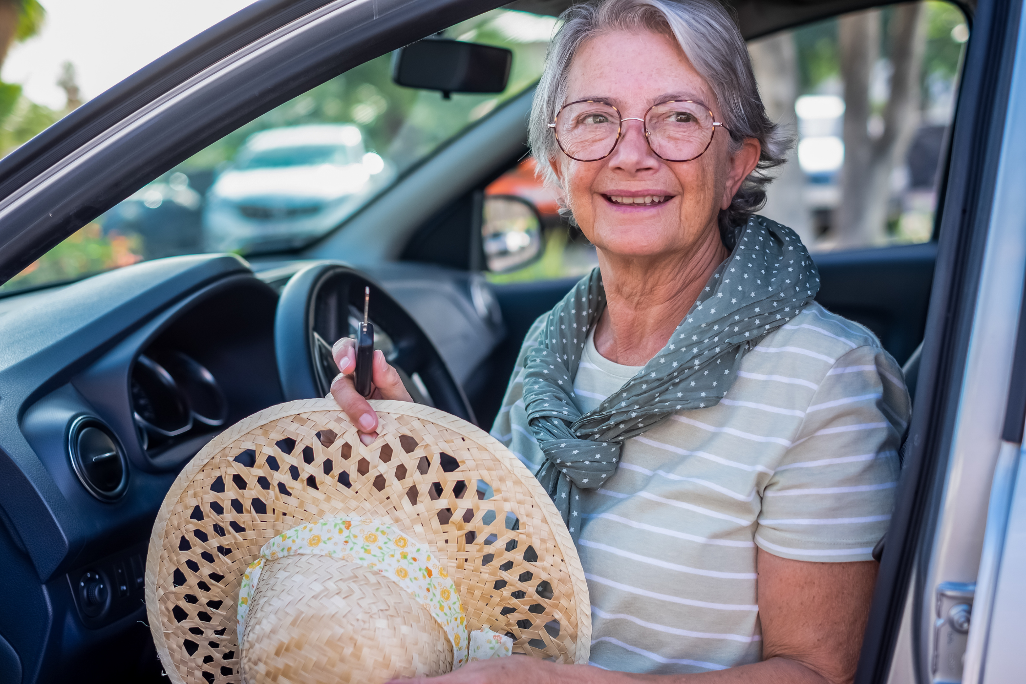 An older woman with glasses and a gray scarf sits in a car holding a straw hat and smiling.