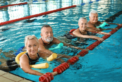 Four smiling older adults in a swimming pool, using foam noodles for support, hold onto the lane divider.