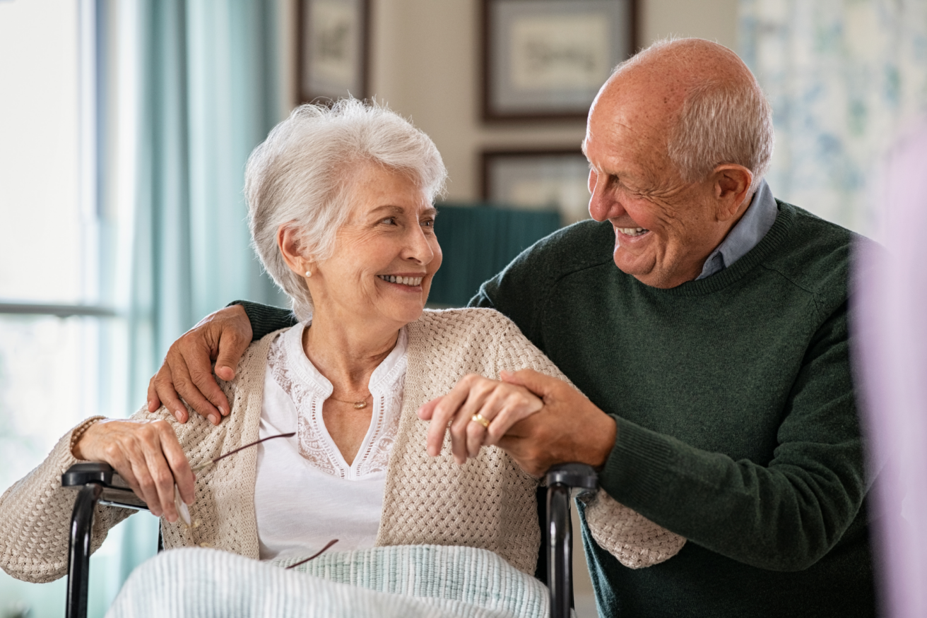 An elderly woman in a wheelchair and an elderly man are smiling and holding hands, sitting close in a well-lit room. The woman holds her glasses in her other hand.