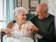 An elderly woman in a wheelchair and an elderly man are smiling and holding hands, sitting close in a well-lit room. The woman holds her glasses in her other hand.