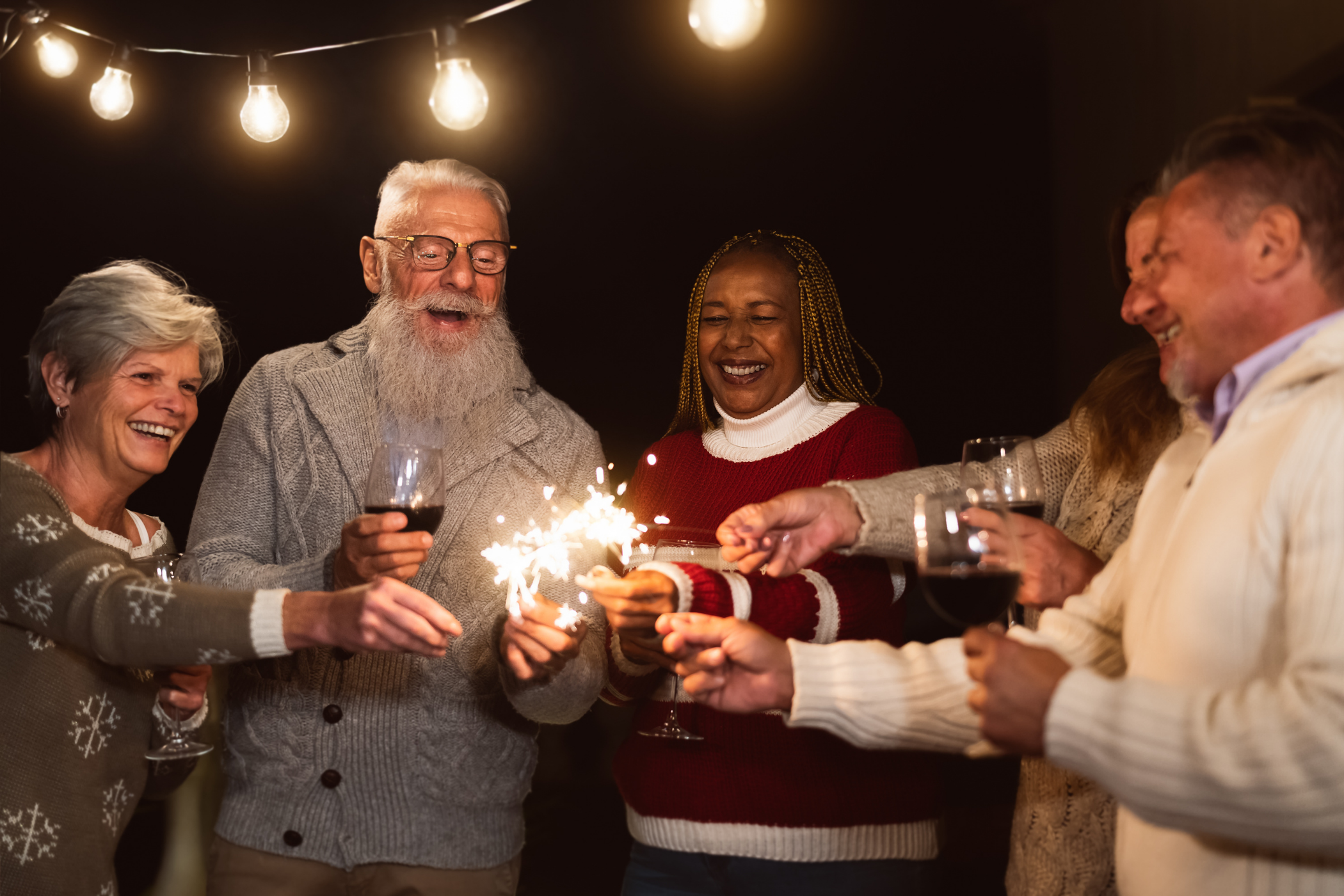 A group of people with glasses and sparklers, smiling and enjoying a celebration under string lights at nighttime.