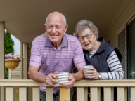 An elderly man and woman stand on a porch, smiling and holding mugs. The man is in a striped polo shirt, and the woman is in a long-sleeved shirt and vest.