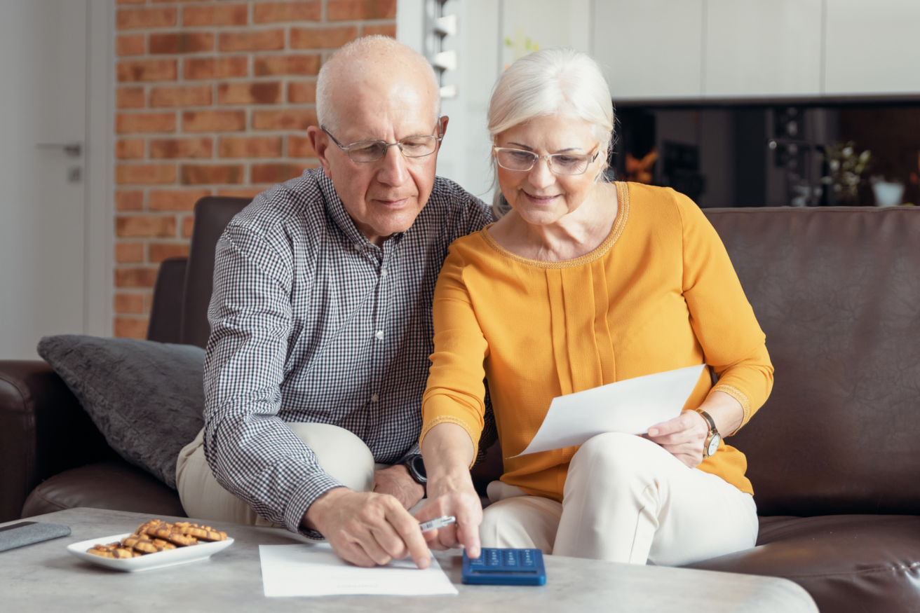 Elderly couple sitting on a couch, reviewing documents and using a calculator. A plate of cookies is on the table in front of them.