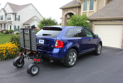 A blue SUV is parked in a driveway with a metal cargo carrier attached to its rear, showing a residential neighborhood in the background.