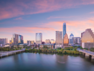 A city skyline at sunrise featuring tall buildings and a calm river in the foreground with a bridge crossing it.