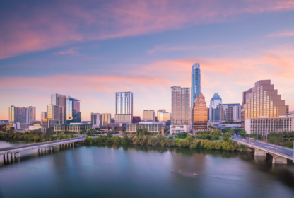 A city skyline at sunrise featuring tall buildings and a calm river in the foreground with a bridge crossing it.