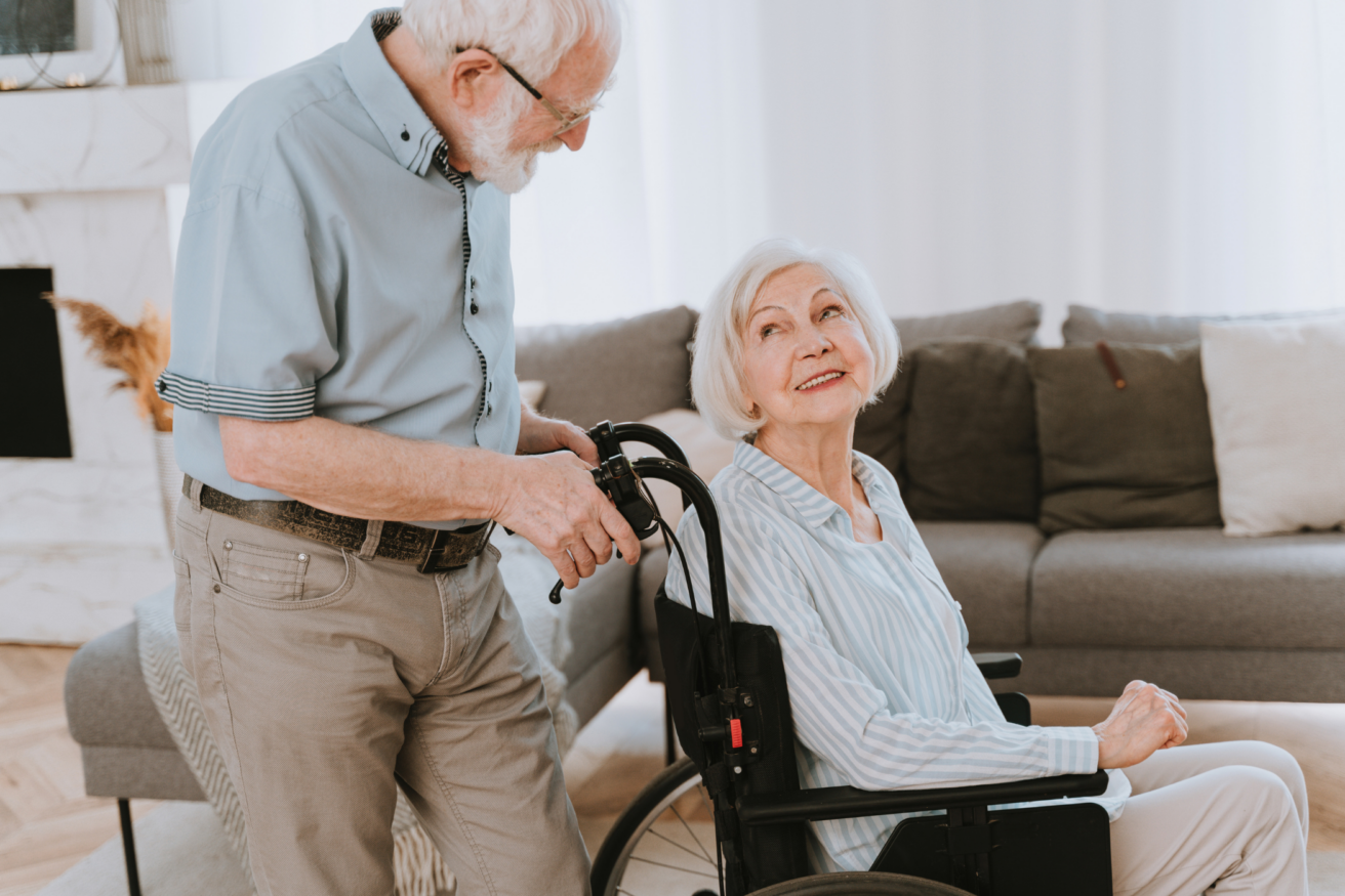 An elderly woman in a wheelchair looks up at an elderly man standing beside her in a cozy living room setting.