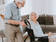 An elderly woman in a wheelchair looks up at an elderly man standing beside her in a cozy living room setting.