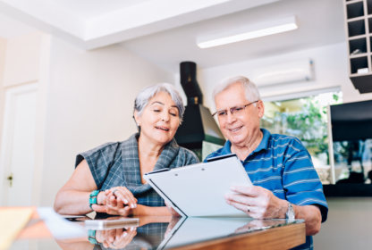 An elderly couple sits at a table in a modern kitchen, looking at a document together. The woman, wearing a gray top, smiles while the man, in a blue striped shirt, reads intently.