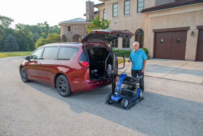 An elderly man standing next to an open minivan, assisted by a mobility scooter lift positioned at the rear of the vehicle.