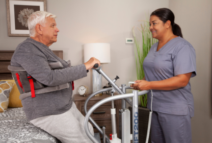 An elderly man using a patient lift is assisted by a nurse in a bedroom setting.