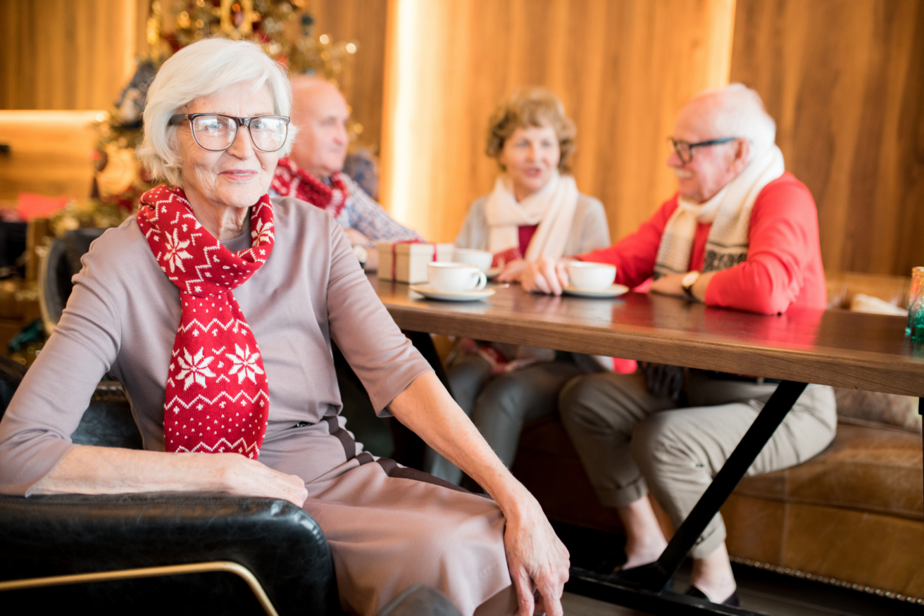 Elderly people in a cozy setting, the focus is on a woman wearing glasses and a red festive scarf, sitting at a table with three others in the background, having a conversation over tea or coffee.