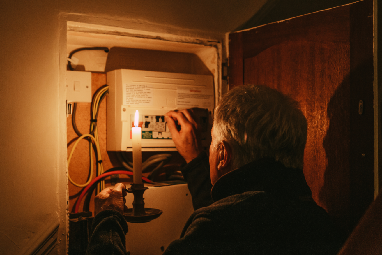 A person holding a lit candle examines an electrical panel inside a cabinet.