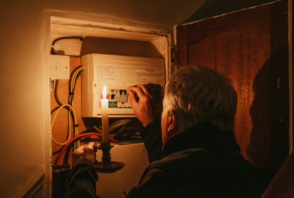 A person holding a lit candle examines an electrical panel inside a cabinet.