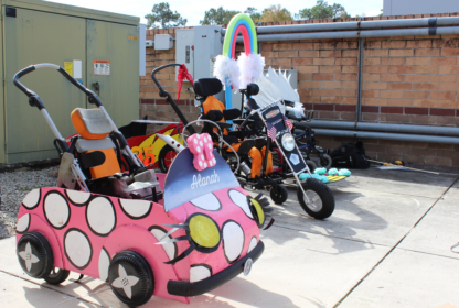 Three decorated children's wheelchairs are parked outside a building: one pink with polka dots, another black with an American flag, and a third with a rainbow and white wings.