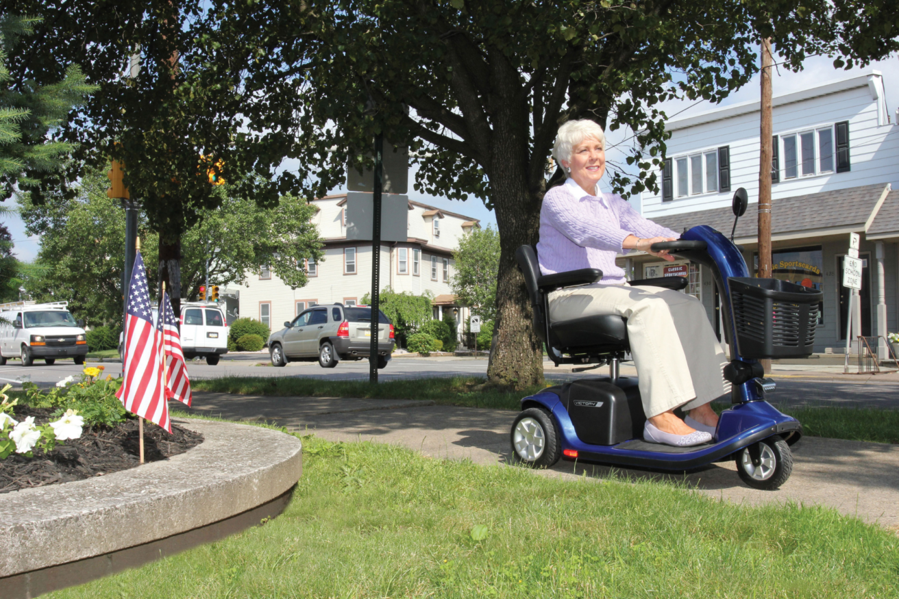 An elderly woman rides a blue mobility scooter on a sidewalk near a tree in a suburban neighborhood. An American flag is planted in a nearby flower bed.