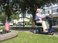 An elderly woman rides a blue mobility scooter on a sidewalk near a tree in a suburban neighborhood. An American flag is planted in a nearby flower bed.