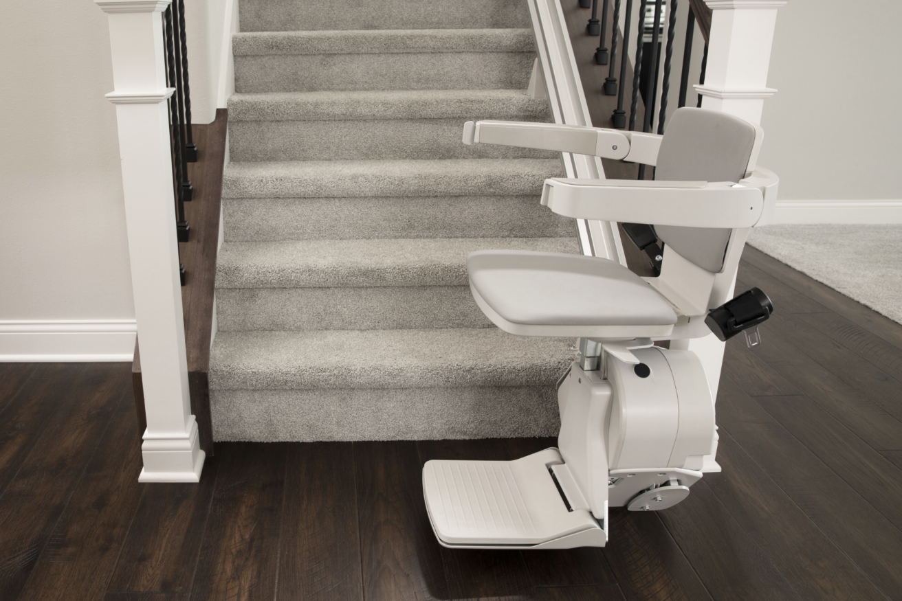 A motorized stair lift installed along the side of a carpeted staircase in a home with dark hardwood floors and white walls.