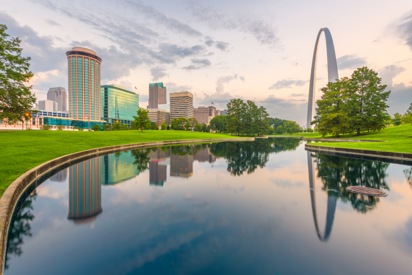 Skyline reflecting in a calm pond at sunrise with the Gateway Arch in the background, surrounded by green trees and buildings.