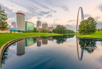 Skyline reflecting in a calm pond at sunrise with the Gateway Arch in the background, surrounded by green trees and buildings.