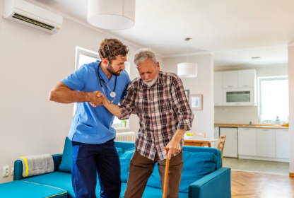 A healthcare professional assists an elderly man with a cane in a living room setting.